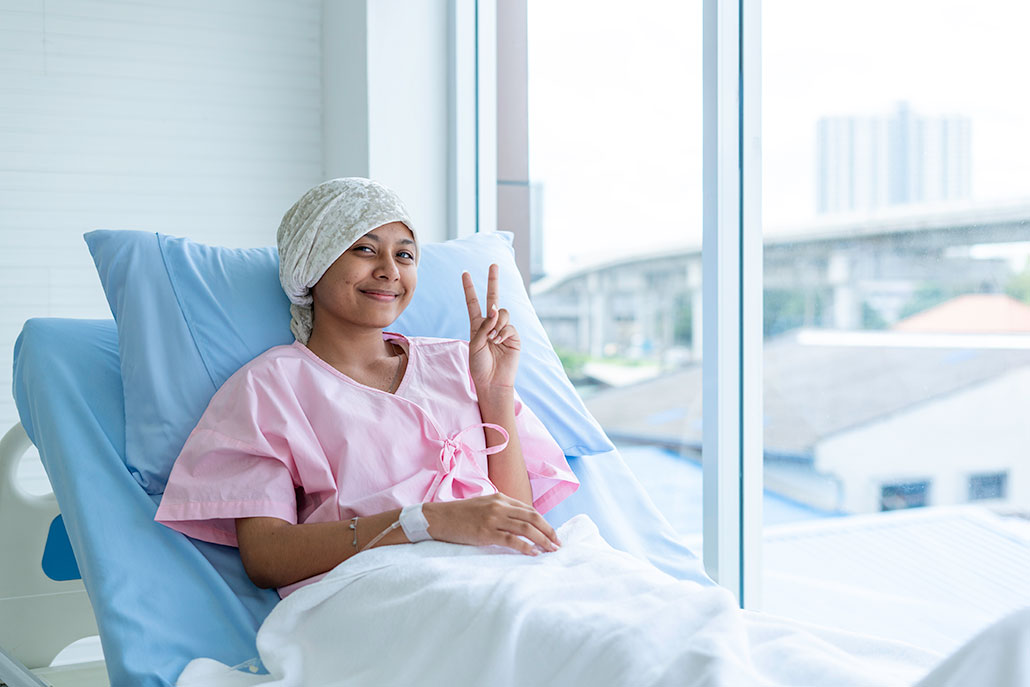 a young woman with light brown skin is sitting in a hospital bed and smiling for the camera. She's making a V sign with her fingers. She is wearing a pink hospital gown and a white headwrap. She is also covered from the waist down by a blanket.