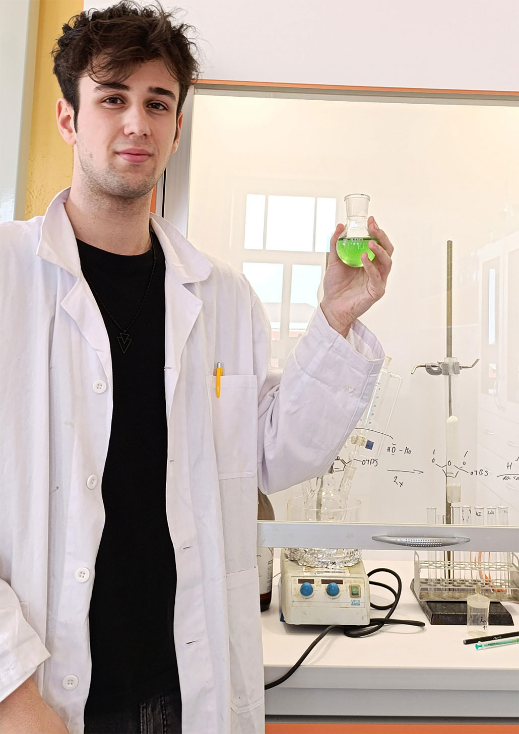 a photo of Kovalčík, a young man with pale skin and dark hair, wearing a lab coat and holding a small glass vial of green liquid in front of a desk set up with organic chemistry lab equipment  for drug synthesis