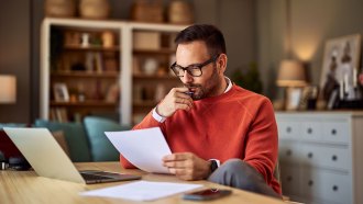 a photo of a man sitting at a desk and reading a piece of paper thoughtfully, with his hand to his face. He's wearing an orange sweater, black rimmed glasses and has pale skin and a short hair cut. Also on the desk is a laptop, another paper, and a phone.