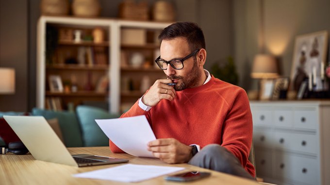 a photo of a man sitting at a desk and reading a piece of paper thoughtfully, with his hand to his face. He's wearing an orange sweater, black rimmed glasses and has pale skin and a short hair cut. Also on the desk is a laptop, another paper, and a phone.