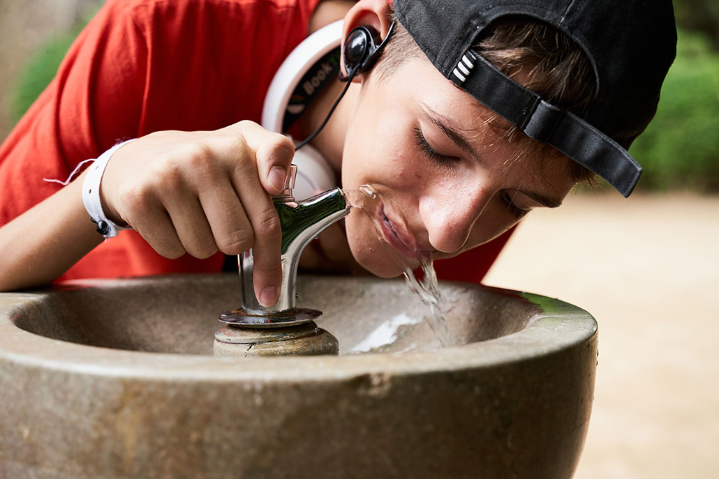 a young man wearing a baseball cap backwards beding over a water fountain to drinik