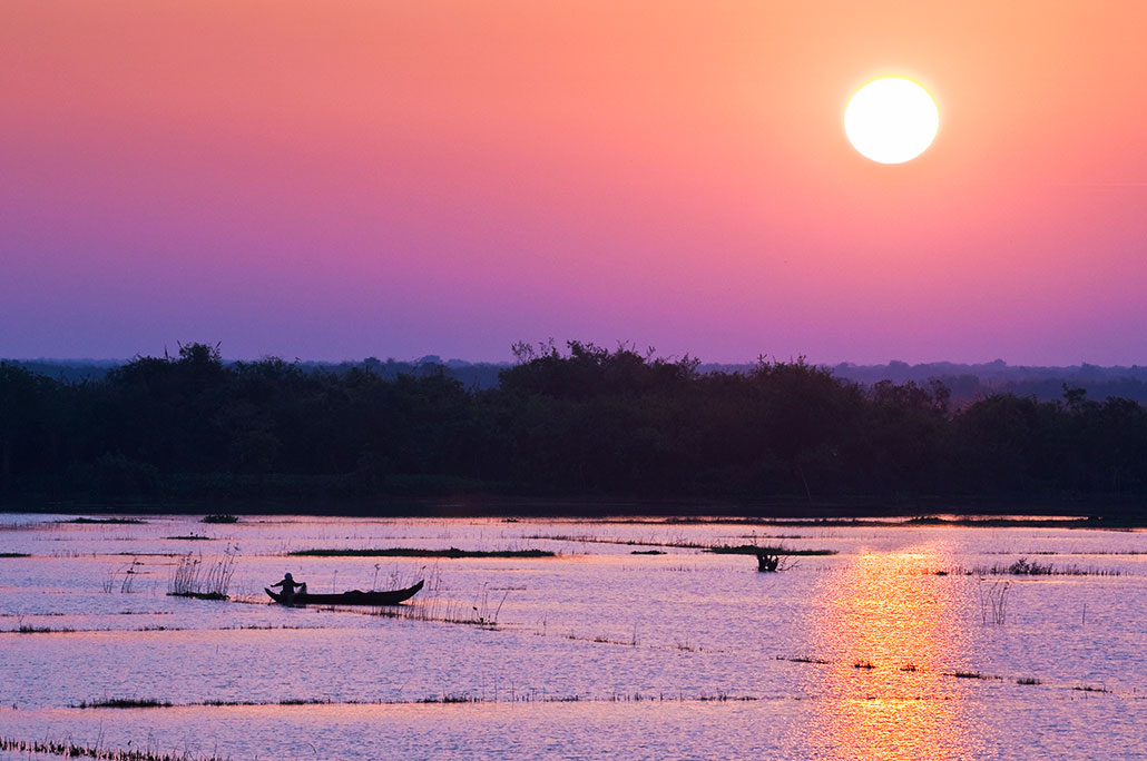 a pink and purple sunset over Tonle Sap Lake, with a person fishing off a boat with a net