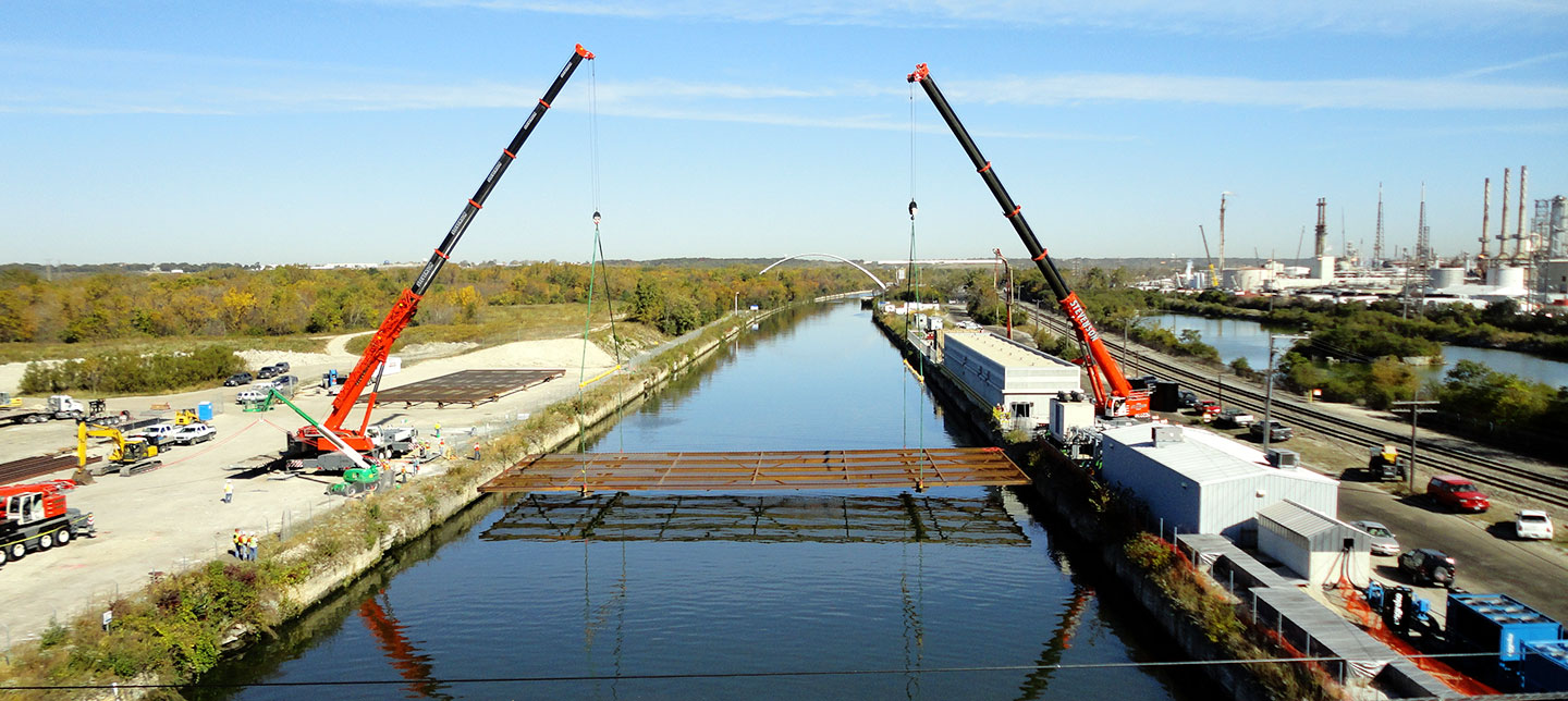 a photo showing two cranes lowering a large rusty metal grate into a canal
