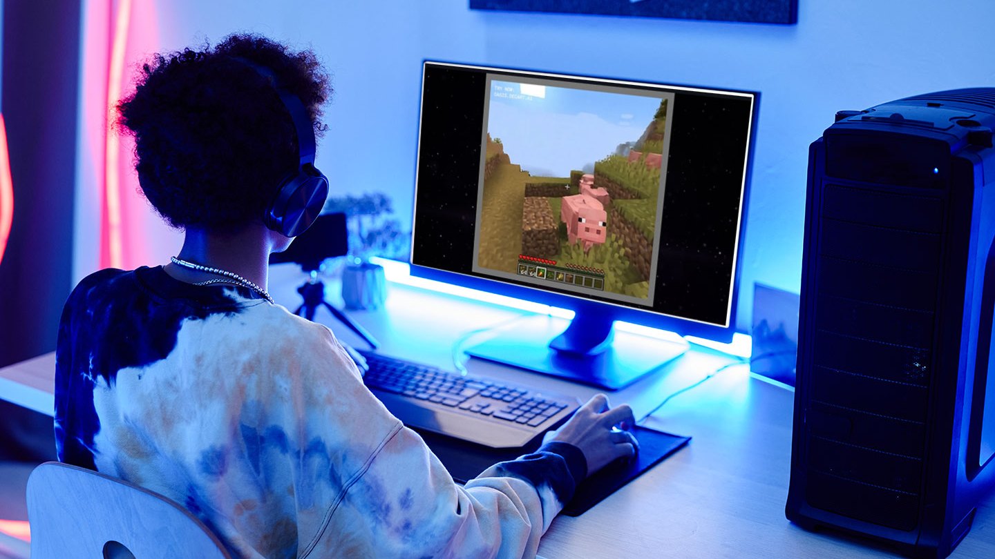 a teen sits in front of a computer looking at a screen with a video game scene AI-generated by Oasis, featuring a Minecraft-like pig. The teen is wearing a black, white and cream tie-dyed hoodie. They have dark brown curly hair and dark skin. Although the room is dark, there are rgb lights behind the monitor and along the wall illuminating the gaming space.
