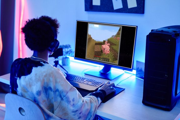 a teen sits in front of a computer looking at a screen with a video game scene AI-generated by Oasis, featuring a Minecraft-like pig. The teen is wearing a black, white and cream tie-dyed hoodie. They have dark brown curly hair and dark skin. Although the room is dark, there are rgb lights behind the monitor and along the wall illuminating the gaming space.