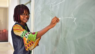 Angela Tabiri at a green chalkboard. She has brown skin, her hair is styled in shoulder length braids and she wears a colorful dress. She also has glasses