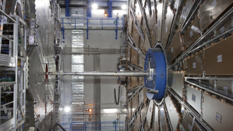 The inside of the not-fully-constructed ATLAS detector has large metallic arrays of panels ATLAS connected by a long rod. The detector searches for leptons and other elementary particles.
