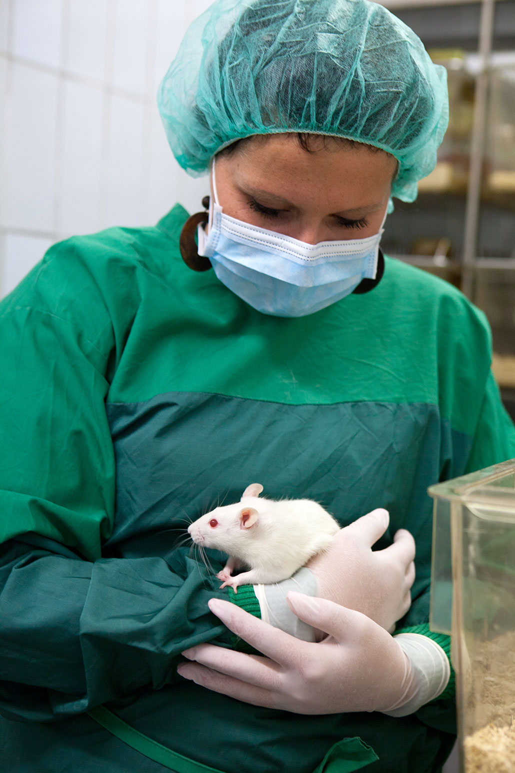 a woman in green lab gear and a surgical mask holds a white rat