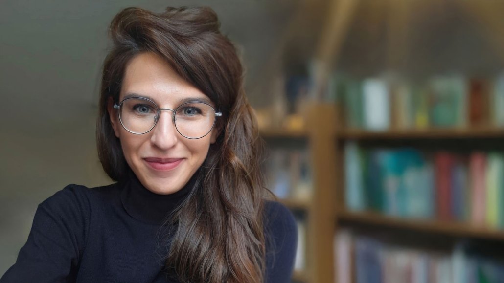  Katarzyna Nowaczyk-Basińska, who researches grief bots, stands in front of a filled bookshelf. She is a white woman with long brown hair and round glasses.