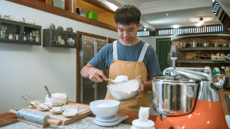 a teen boy measures flour into a bowl on a table filled with baking supplies to make bread