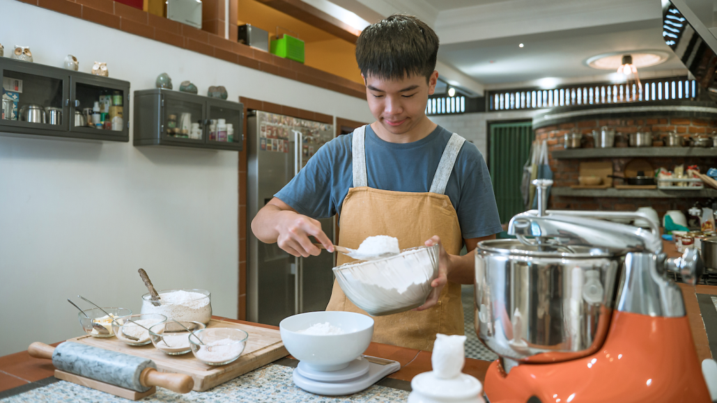 a teen boy measures flour into a bowl on a table filled with baking supplies to make bread