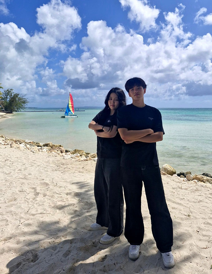 two teens stand back to back with arms crossed on a white sand beach, under a sunny sky