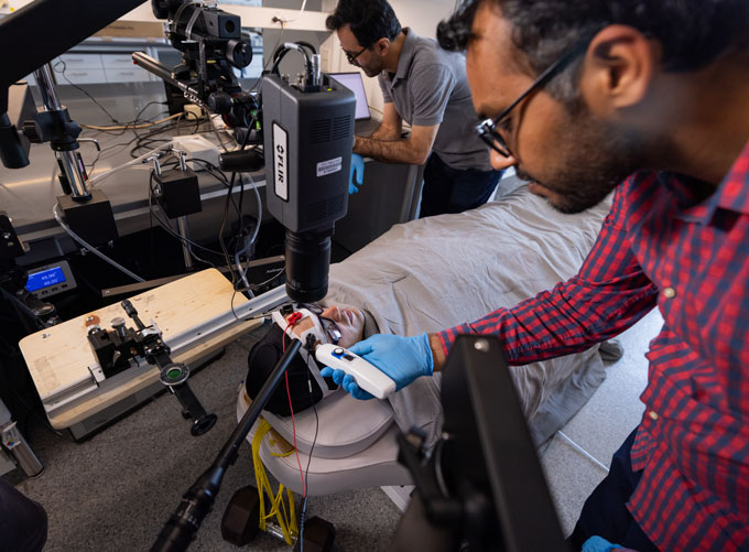 A person wrapped in a specialized suit and covered in a blanket lies with only his head visible. He's surrounded by equipment, and two colleagues, to measure his sweat.