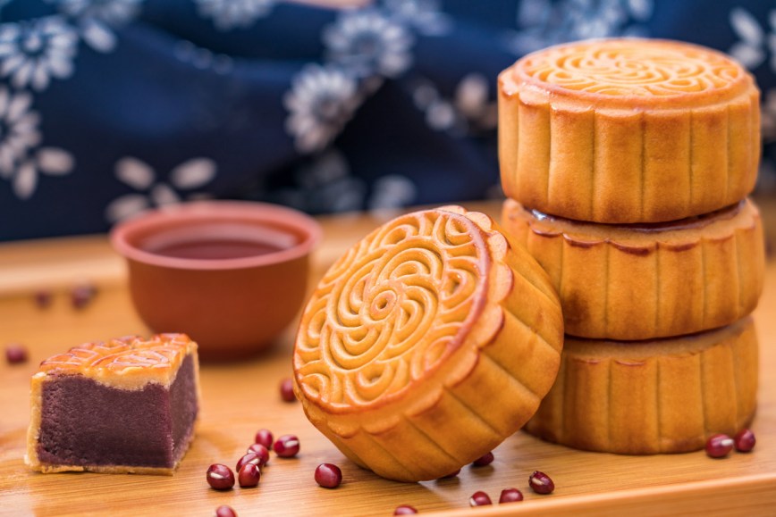 A stack of brown mooncakes sit on a wooden table. Red beans are scattered nearby.