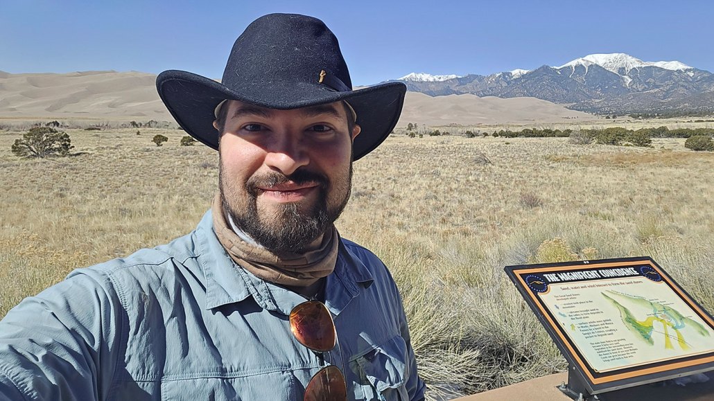 Biologist Juan Manuel Vazquez stands in front of a grassland. Mountains loom behind him in the distance. He's a Latino man wearing a wide brimmed black hat and blue shirt.
