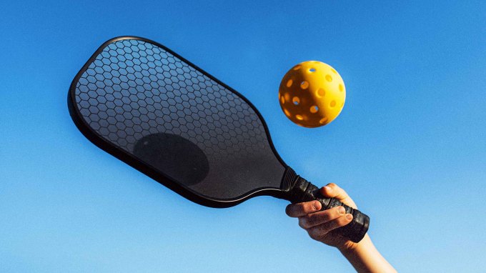 a photo of a hand holding a pickleball paddle about to hit a yellow pickleball