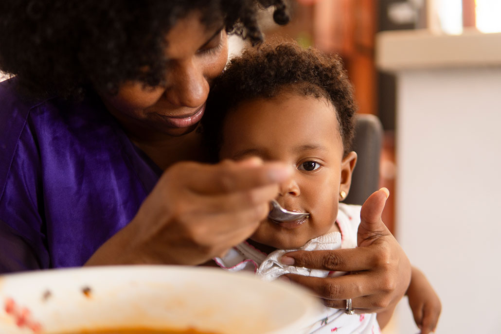 a young baby with brown skin and fluffy dark hair is sitting in a woman's lap and being fed baby food