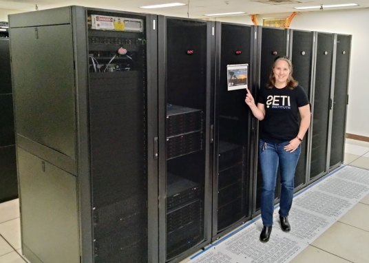Chenoa Tremblay stands in front of a row of supercomputer servers. She is a white woman with brown, shoulder-length hair. She is wearing blue jeans and a black T-shirt with "SETI" spelled out on the front.