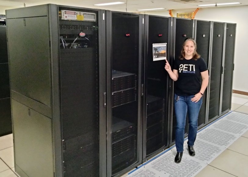Chenoa Tremblay stands in front of a row of supercomputer servers. She is a white woman with brown, shoulder-length hair. She is wearing blue jeans and a black T-shirt with "SETI" spelled out on the front.