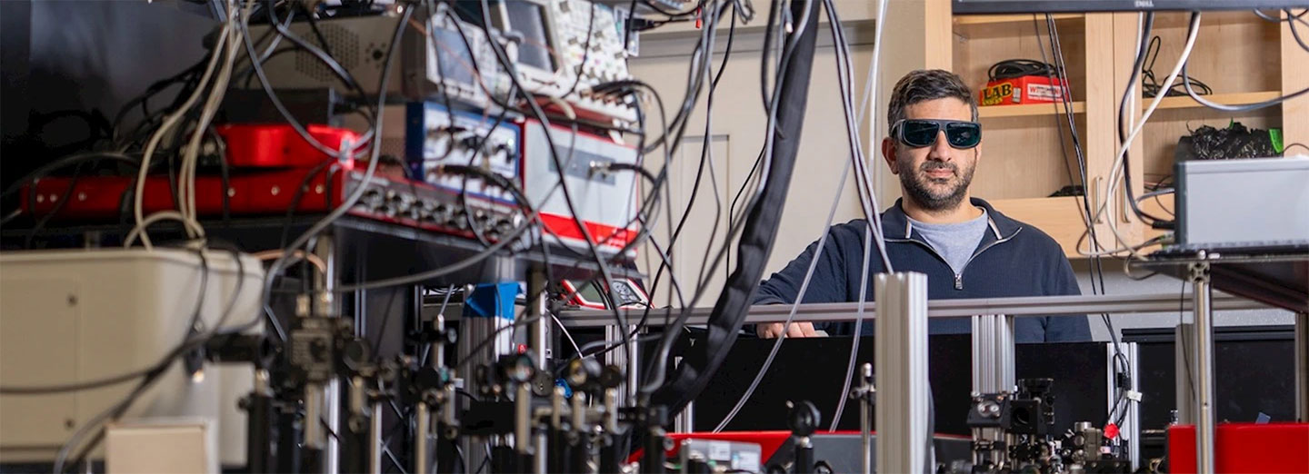 a photo of Elad Harel sitting in a lab behind a lot of equipment. He is wearing dark glasses and listening intently