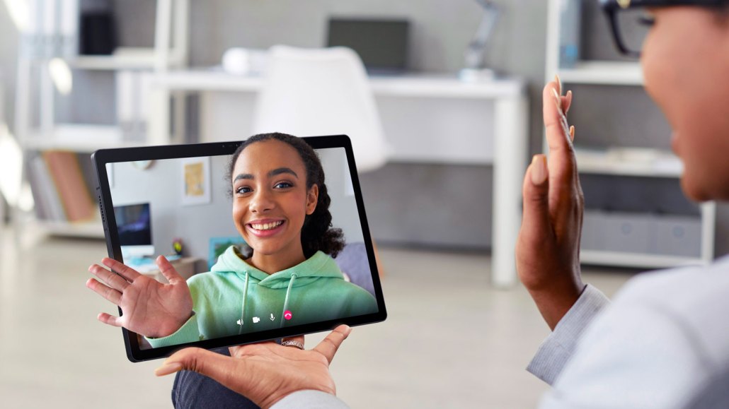 a person seen in profile is wearing black rimmed glasses and waving at their friend on some type of screen technology. The friend seems to pop out of the screen like a hologram.