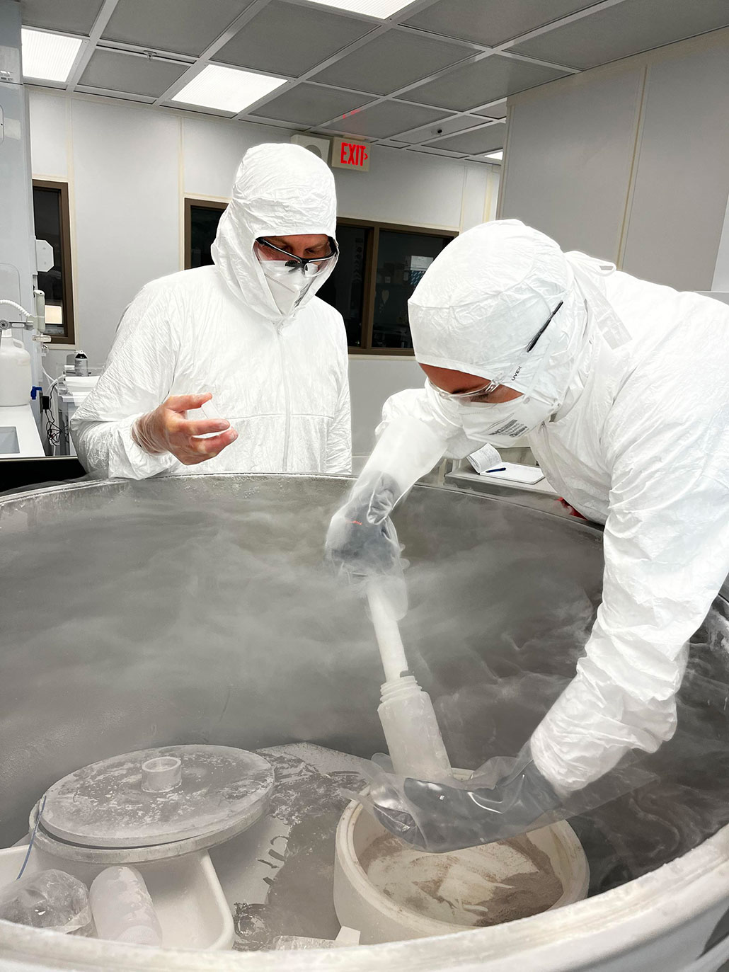 two researchers in white clean suits, masks and goggles are retrieving fecal samples from a large metal container used to keep the samples in a deep freeze.