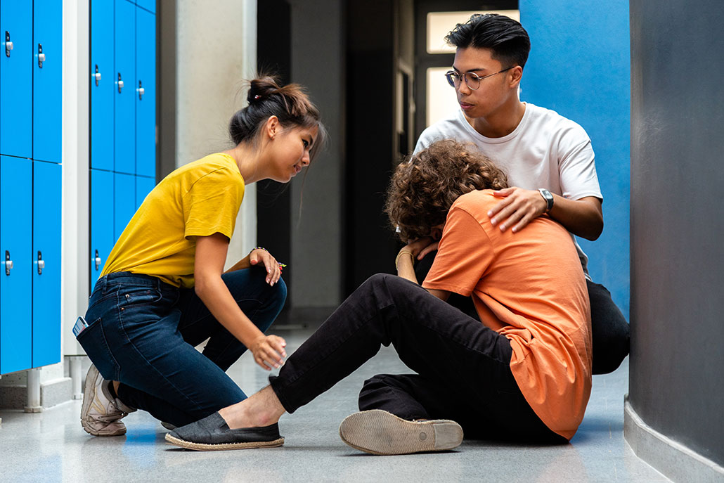 two students comfort another student who is on the floor of a locker room, distraught