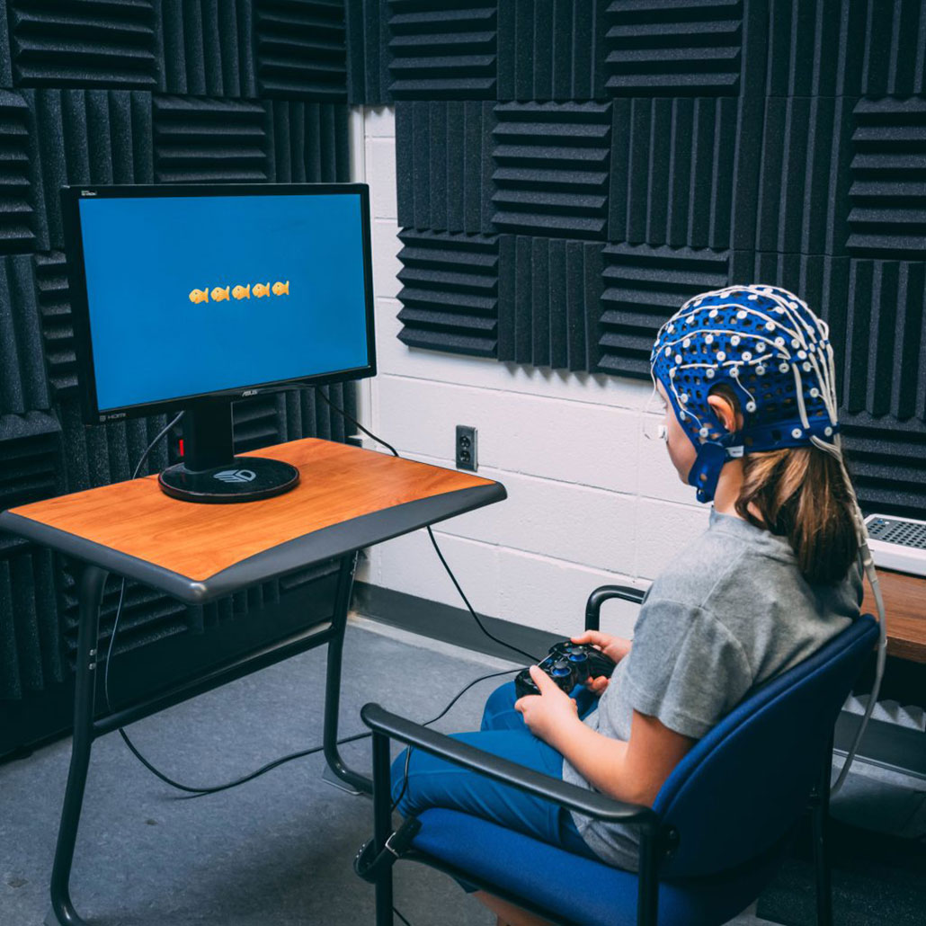 a girl wearing a blue EEG cap sits in front of a monitor with five gold fish on the screen, she's holding a game controller