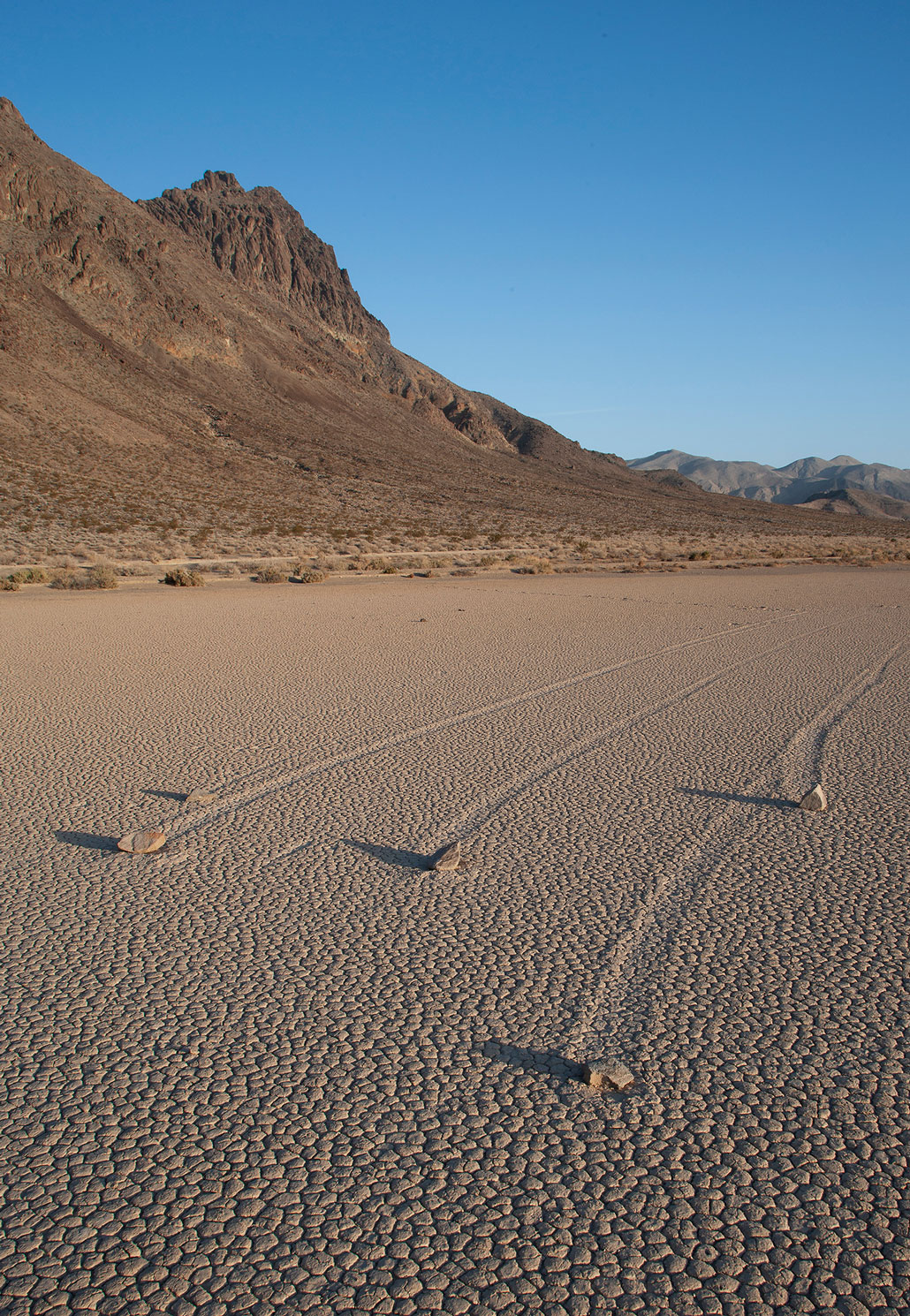 a photo of a barren dirt desert landscape, mountains are in the distance. The sky is bright blue and clear and there are 5 boulders with trails behind them that have all traveled towards the viewer.