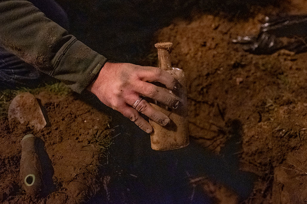 a man's hand caked in red dirt, holding a very old glass bottle near the ground it was unearthed from