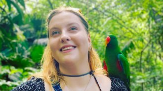 Linguist and languages constructor Margaret Ransdell-Green stands in front of trees with a green parrot on her shoulder. She is a white women with blonde hair and fair skin. She is wearing a black dress with small dots.