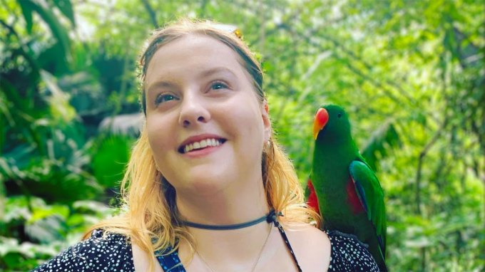 Linguist and languages constructor Margaret Ransdell-Green stands in front of trees with a green parrot on her shoulder. She is a white women with blonde hair and fair skin. She is wearing a black dress with small dots.