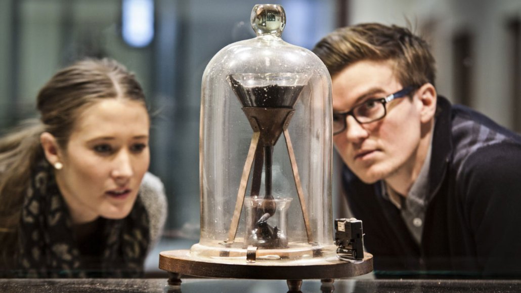 a man and a woman are staring intently at a funnel of pitch under a glass case