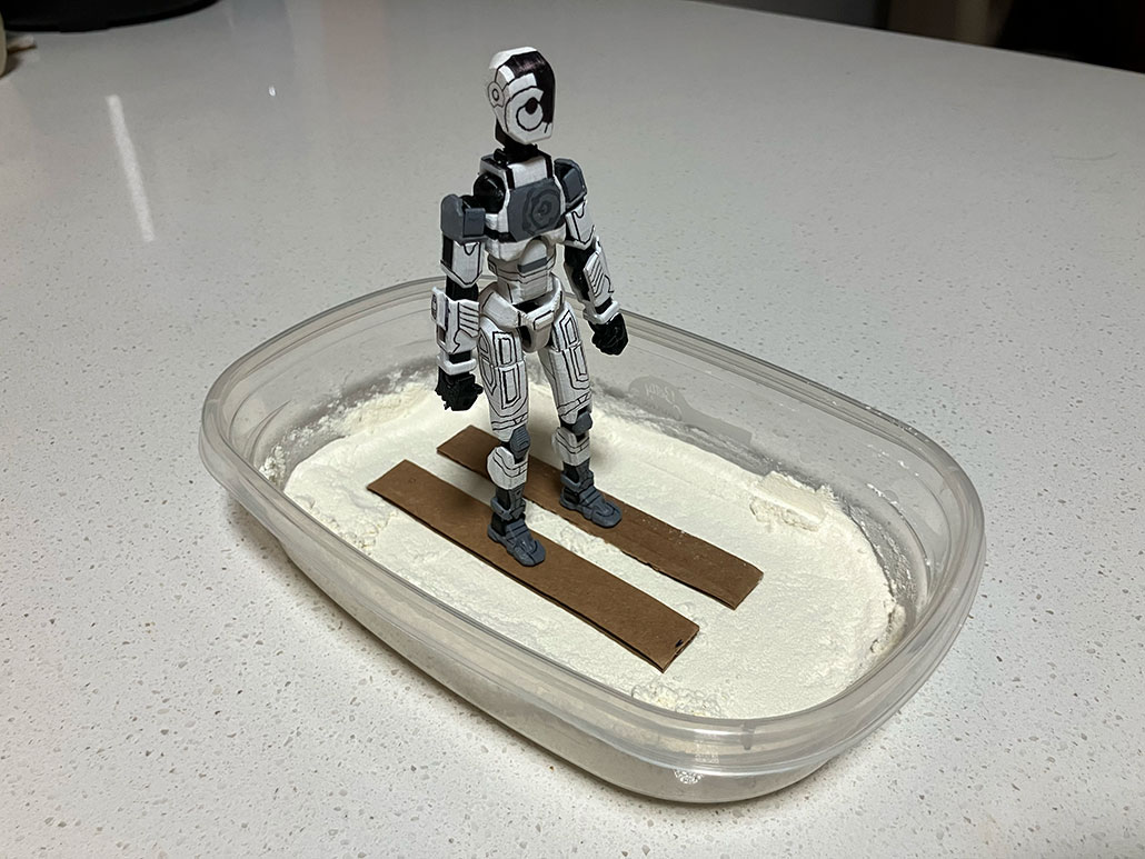an action figure with cardboard skis glued to its feet stands in a container on a layer of flour