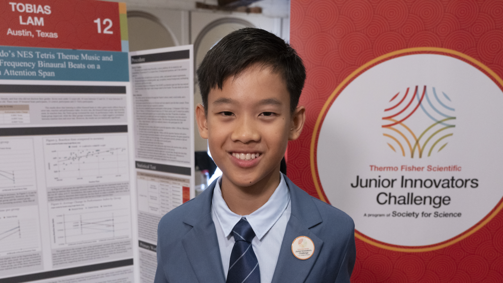 Toby Lam stands in front of a red background and his poster about how sound can affect people's focus during a task.