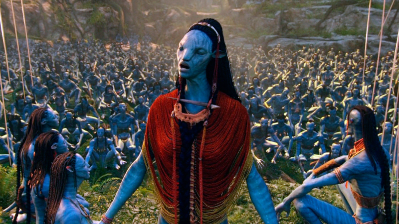 A blue Na'vi humanoid alien from Avatar closes her eyes as she sings in front of a gathered crowd of Na'vi sitting on the ground behind her