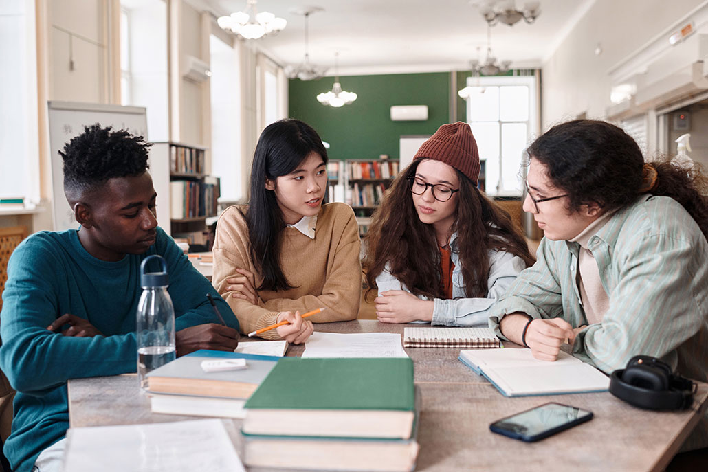 Four young adults sitting around a table and studying together. One of the phone has been place farther away so as not to tempt the studiers.