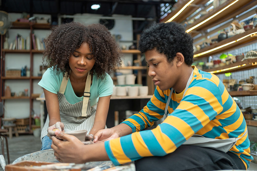 a woman with dark skin and fluffy curly hair is wearing an apron and bending down to help a student shaping a pot on a pottery wheel. They are in a pottery studio.