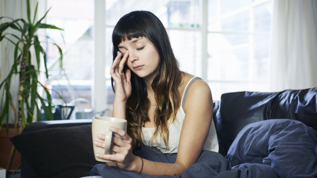 a teen girl sits on a couch holding a mug, sleepily rubbing one of her eyes