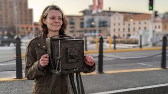 Game designer Susannah Emery stands behind a statue of a camera. She is a white woman with brown eyes and brunette hair. She is wearing glasses and a muted green jacket.