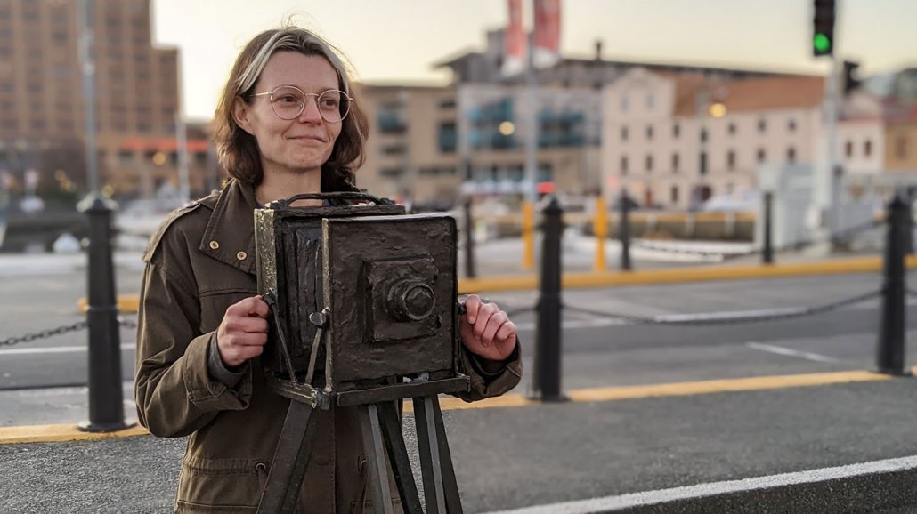Game designer Susannah Emery stands behind a statue of a camera. She is a white woman with brown eyes and brunette hair. She is wearing glasses and a muted green jacket.