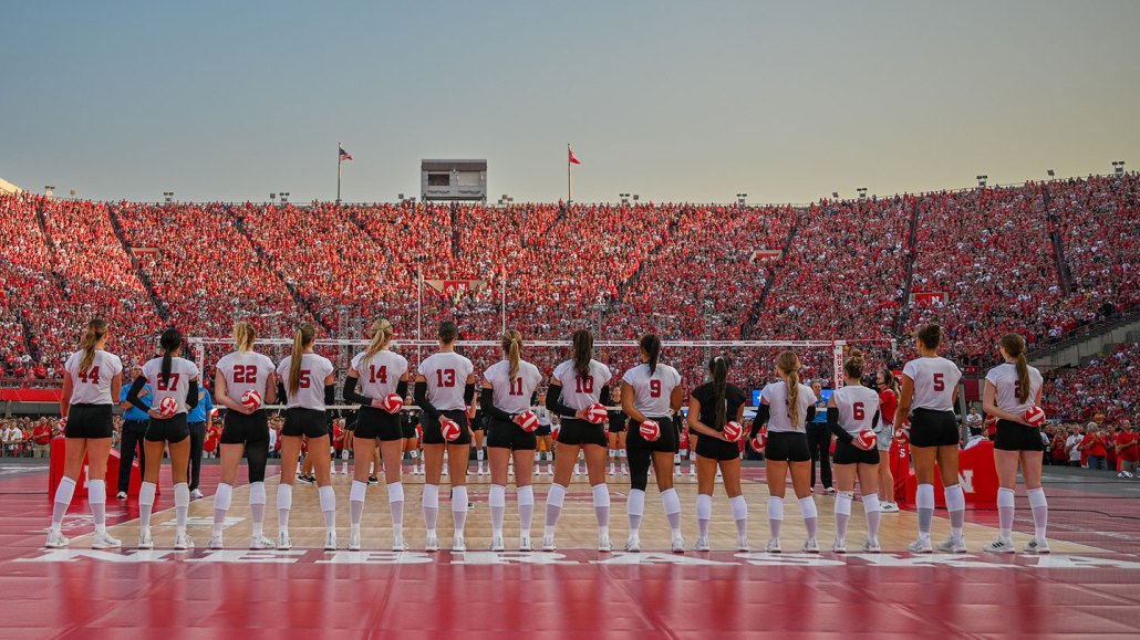 a woman's volleyball team in a full arena facing their audience. The women are standing in a straight line, holding volleyballs behind their backs.