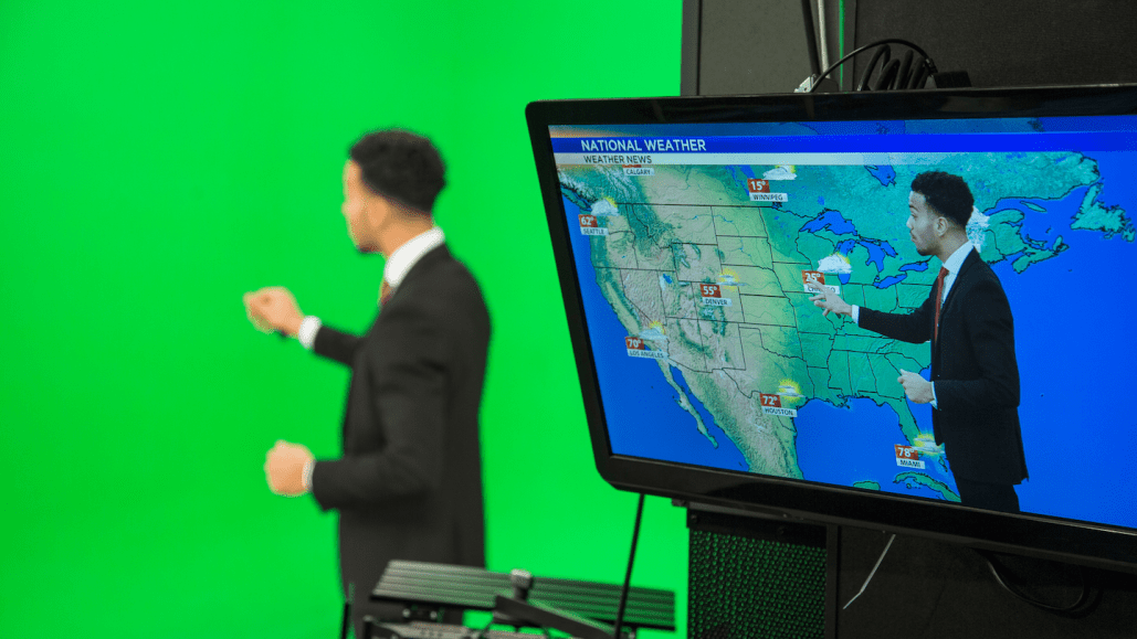 a weatherman stands in front of a green screen, pointing at something; nearby, a TV screen shows what viewers see at home-- the weatherman standing in front of a map of the united states