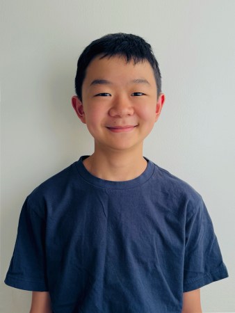 a middle school boy in a blue shirt and standing against a white wall smiles at the camera