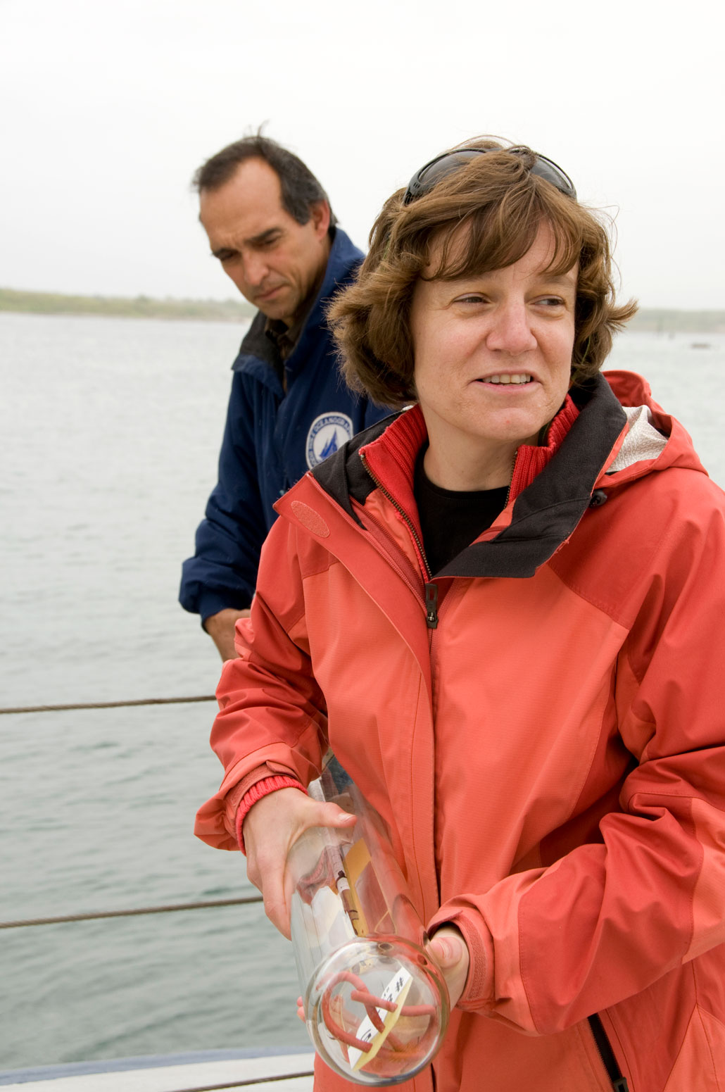 A woman with pale skin and a short brown wavy hairstyle is on a boat. She is holding one end of an ocean science tool. The device appears to be a long glass tub with something inside it at the bottom. There is a man behind her also helping her hold the device.