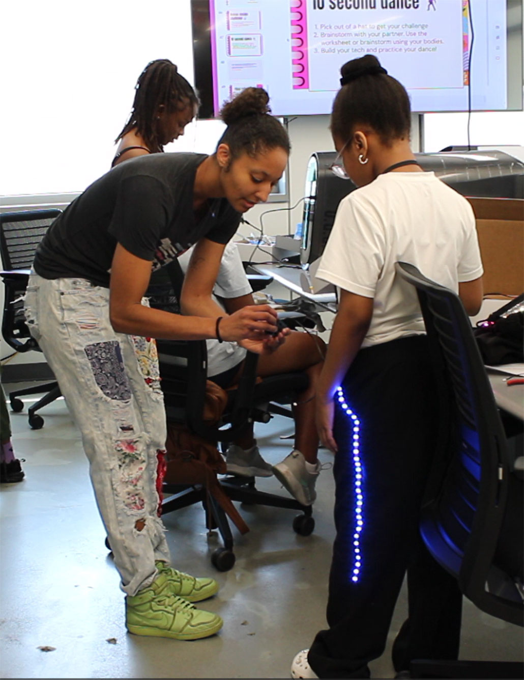 Kayla DesPortes, a brownskinned woman with an updo and jeans with artsy patches on them, is bending over to show the controls for LEDS in a young dancer's pants. They are in a computer lab.