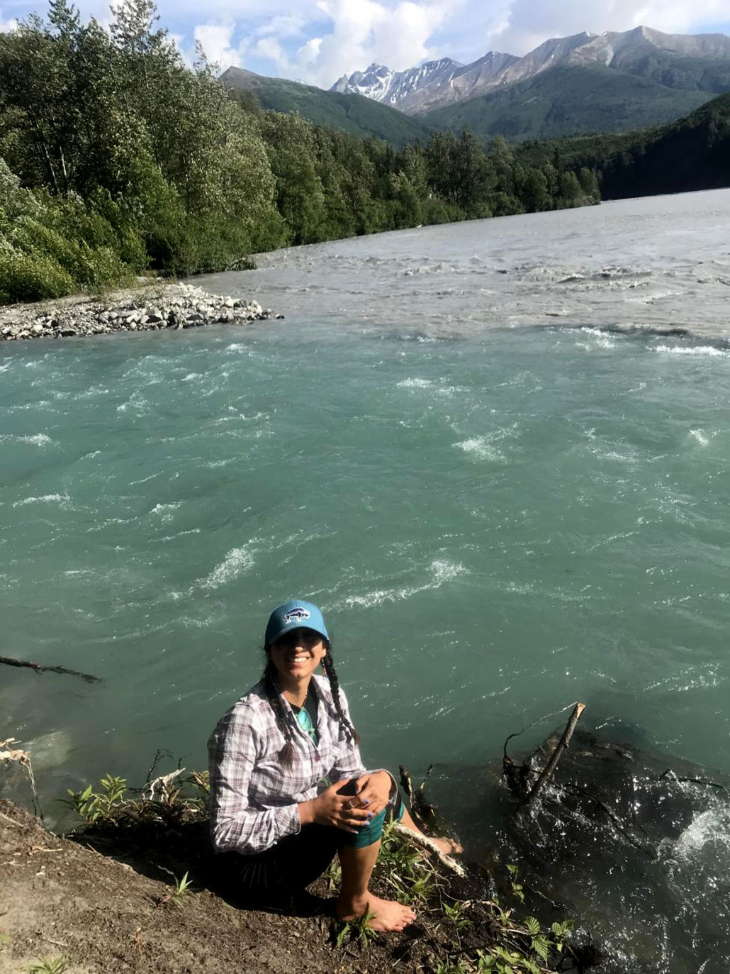 a photo of Lydia Jennings, a woman with a plant shirt, green pants and a blue ball cap. Her long dark hair is in two braids. She is sitting on the bank of a river with a stunning forest and mountain landscape behind her.