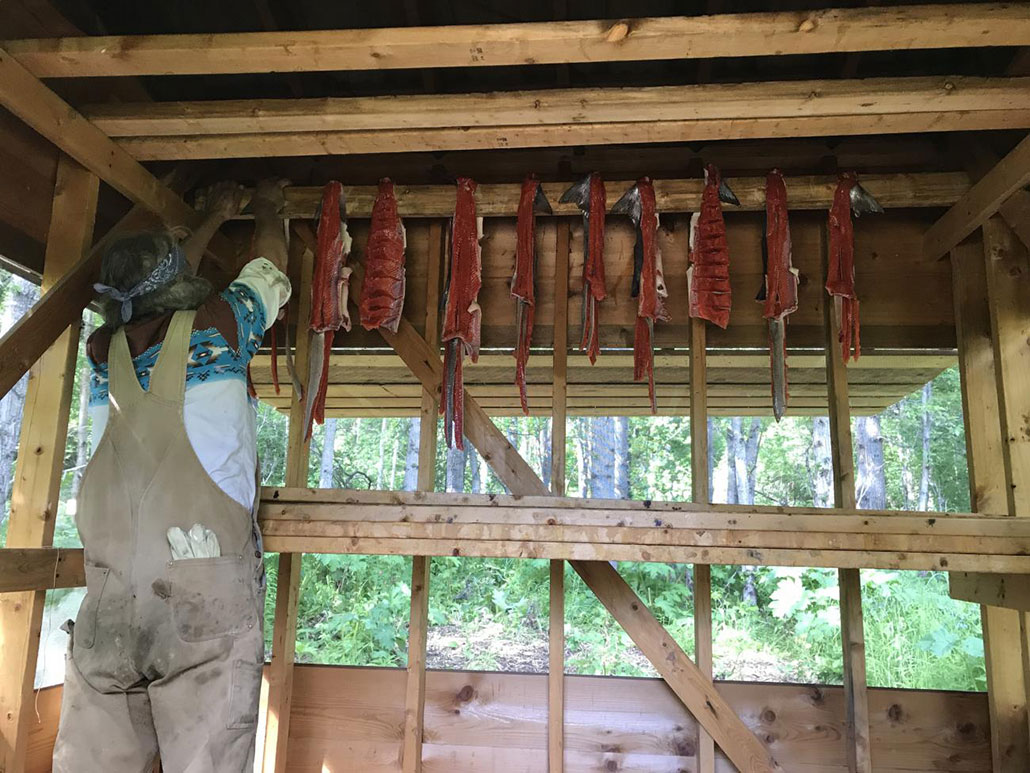 a person standing on a stool hangs salmon to dry in a wooden building that is open on the sides to the elements