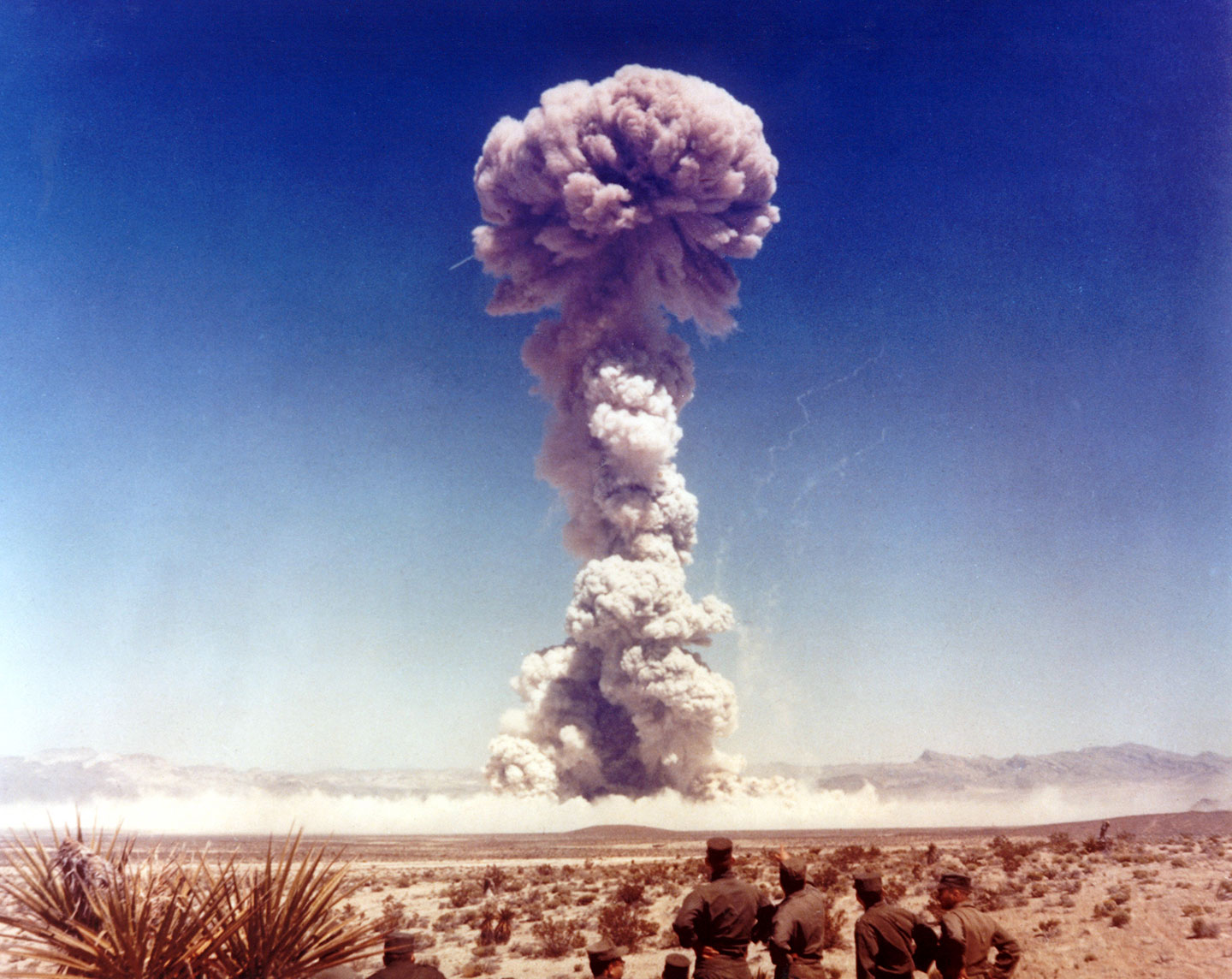 In the distance mushroom cloud reaches high up into the sky over a dry desert. In the foreground several uniformed troops are viewing the nuclear weapon test.