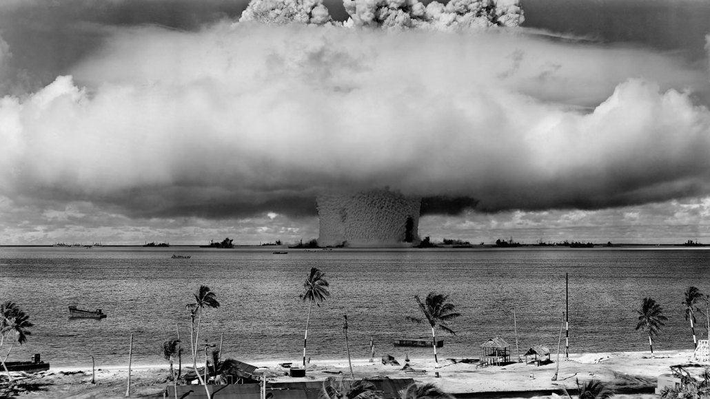 a giant mushroom cloud billows out from Bikini atoll, as seen from a nearby beach